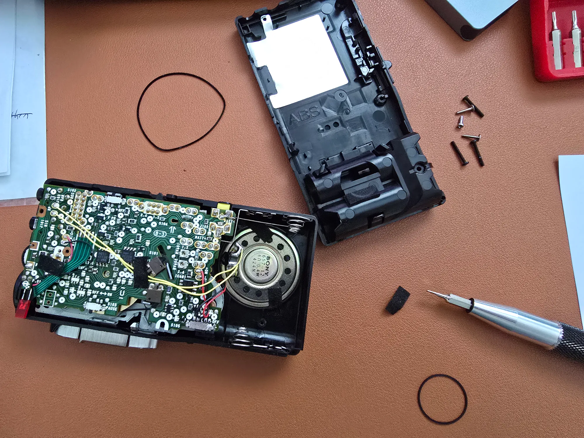 The Sony M-677V microcassette recorder  has the top panel removed, exposing the device's circuit board. There is a speaker device that is taped down with some fuzzy black tape, and from that speaker are several wires that are also taped to the circuit board. The wires are taut giving little clearance to move the circuit board even after the tape is removed from the wires. The picture also has the old, worn belt in the top left corner. The worn belt is not a perfect circle and has two irregularities from where it was pulled taut for years while in storage. In the bottom right corner is another new belt that is a perfect circle without irregularities 