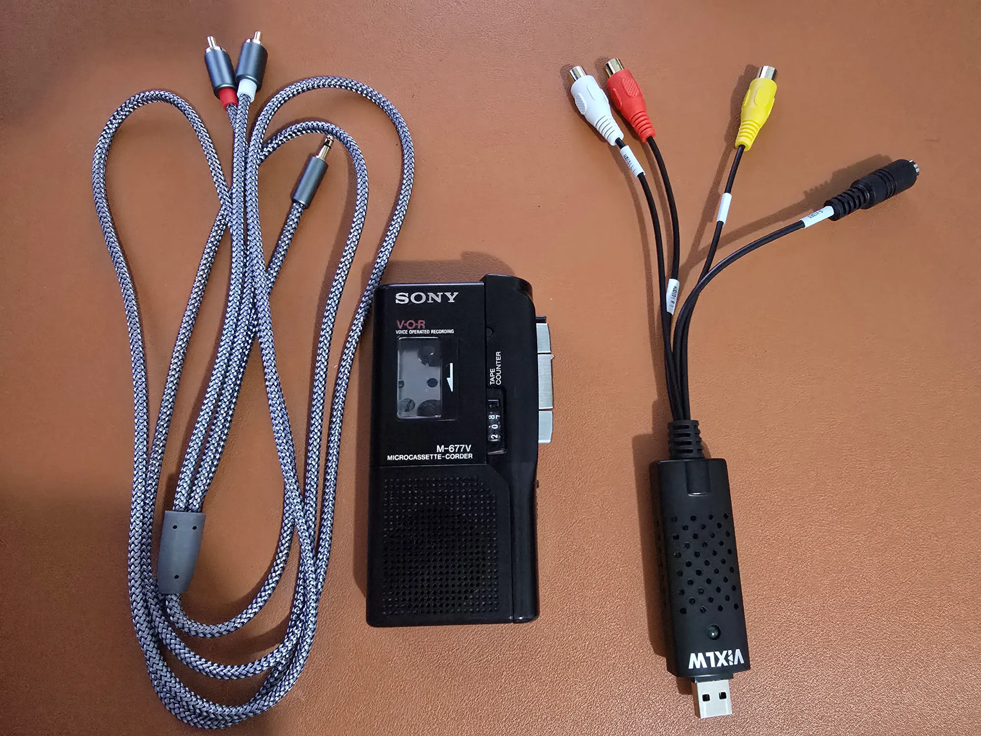 There are three devices on a tan desk-mat. The devices are a 3.5mm audio cable, a microcasette player, and a USB capture card. The devices are separated in this picture and merely side by side